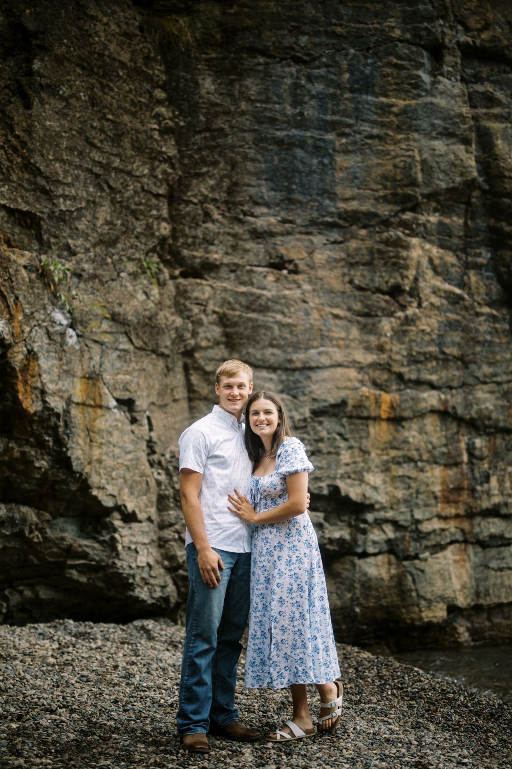 A couple in white and floral dress standing together against a dramatic rocky cliff face backdrop.