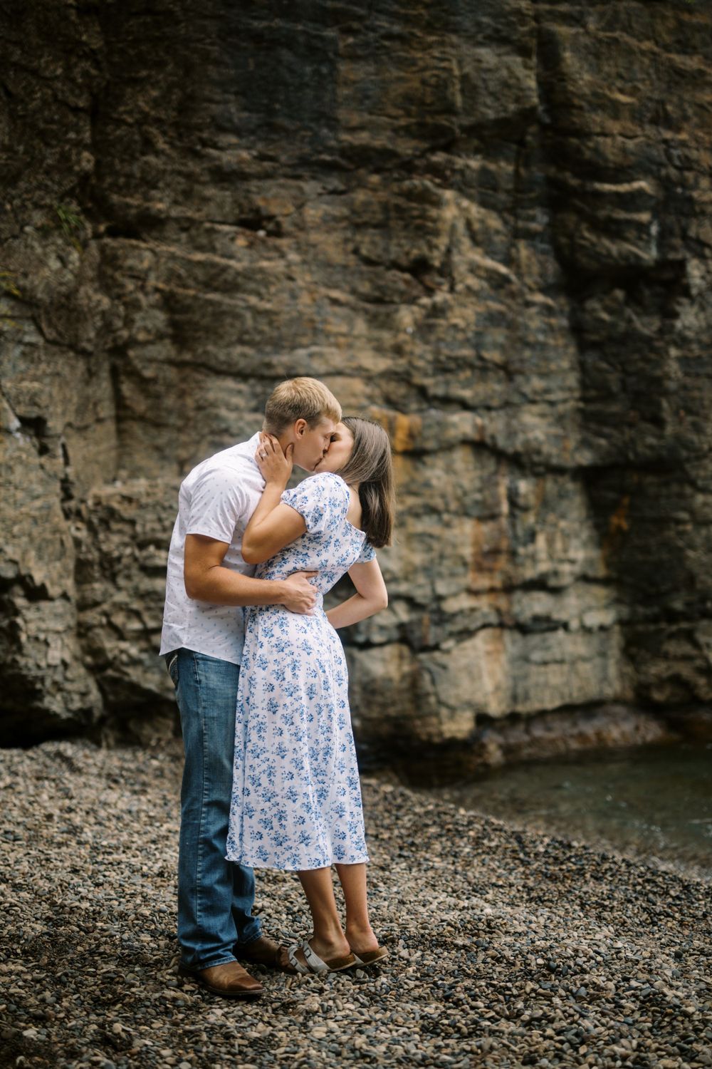 A romantic couple embraces against a dramatic rocky cliff face beside a body of water.