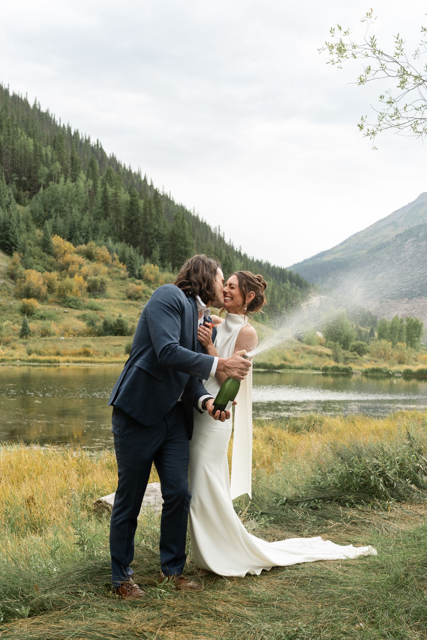 bridal outfits on bride and groom. standing in front of a pond and mountain in colorado. Spraying champagne and sharing a smiling kiss.