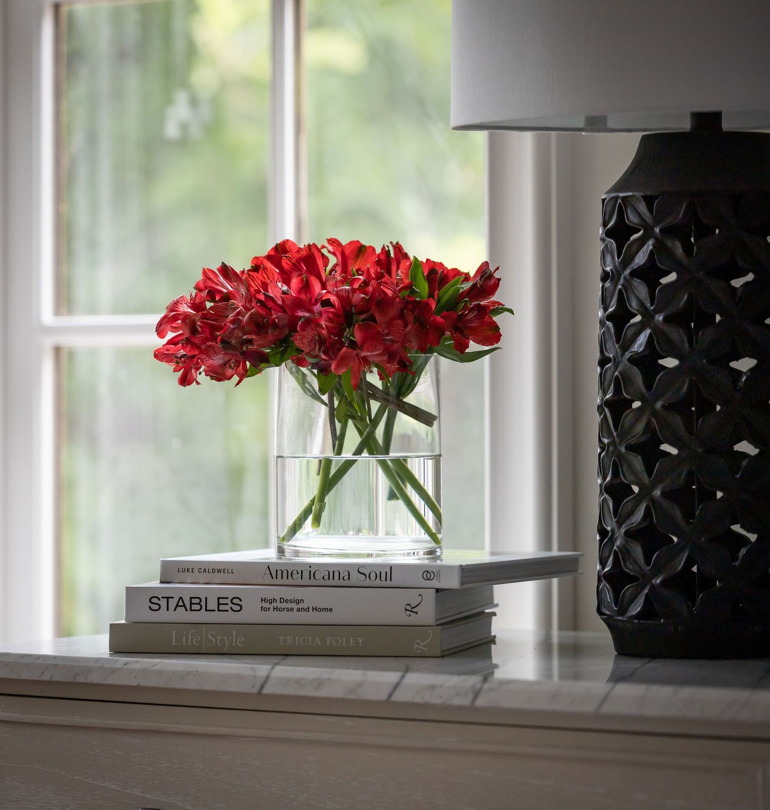 Red flowers in a clear vase with books and black decor on a white windowsill.