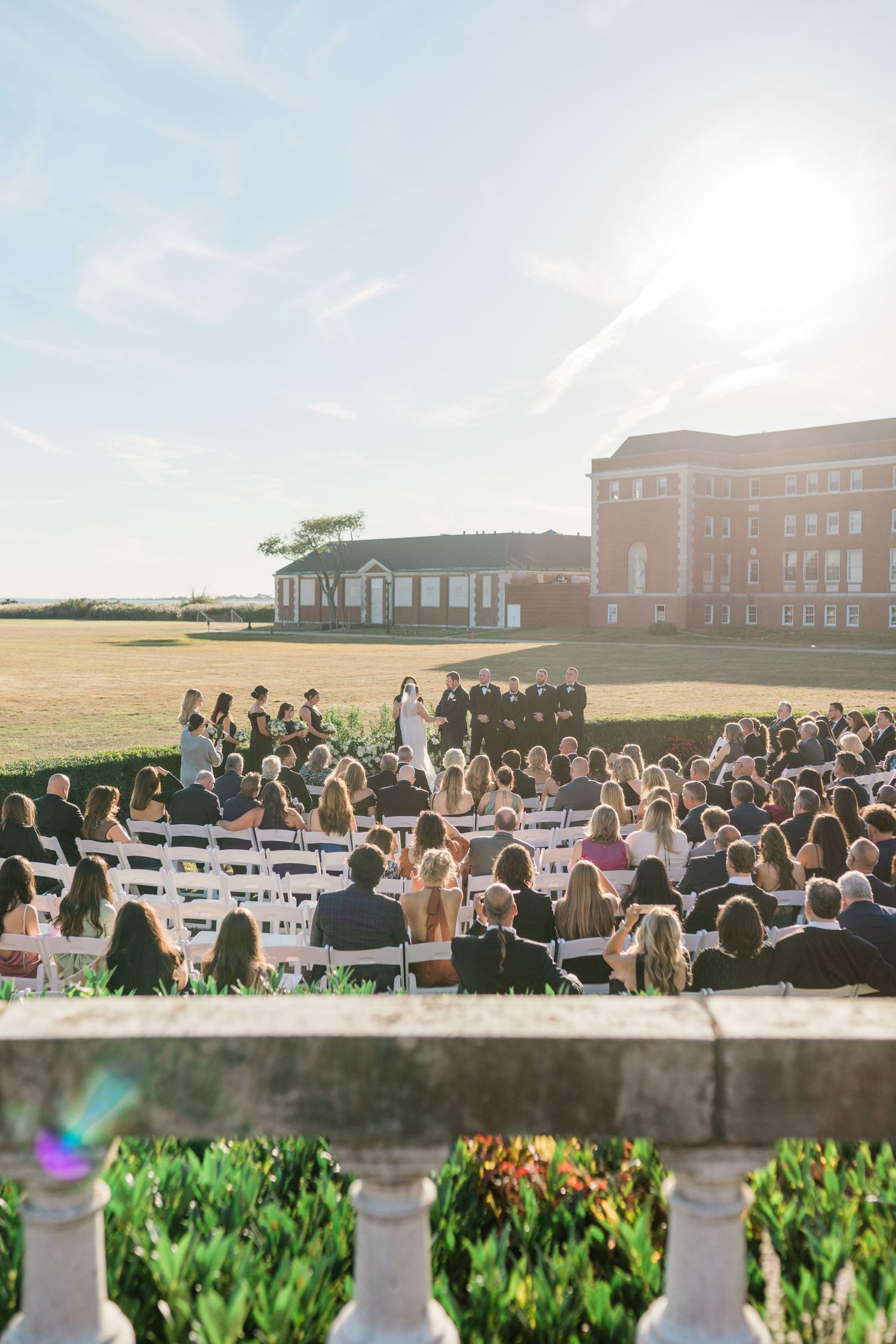 Wedding ceremony guests seated outdoors on a sunny day with brick buildings in the background.