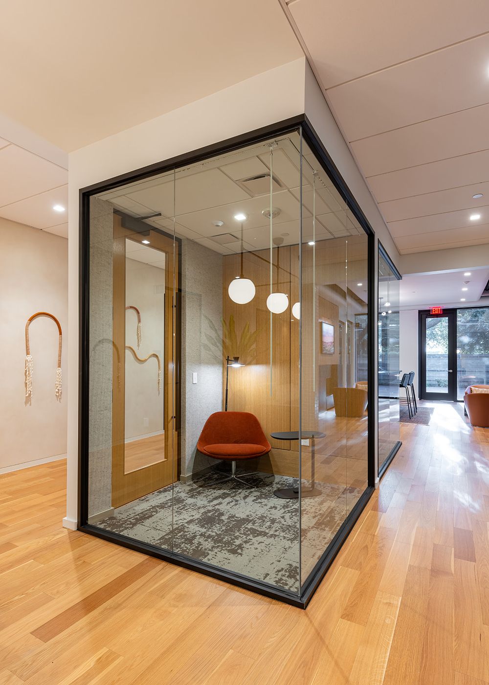 Modern office interior with glass phone booth featuring red chair, pendant lights and hardwood floors.