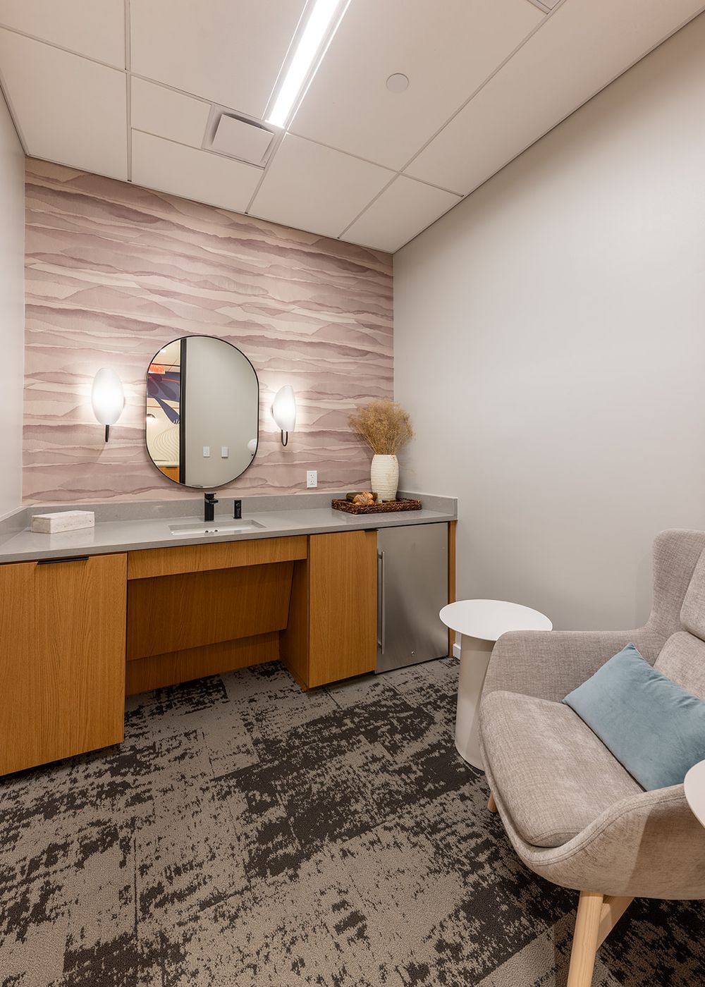 Modern vanity room with oval mirror, wooden cabinets, textured accent wall, and gray upholstered chair on distressed carpet.
