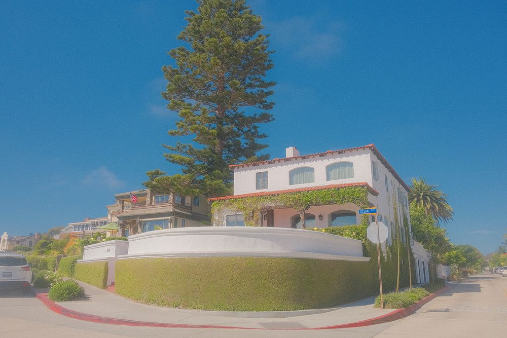 Mediterranean-style white stucco house with curved walls and tall pine tree on sunny hilltop corner lot.