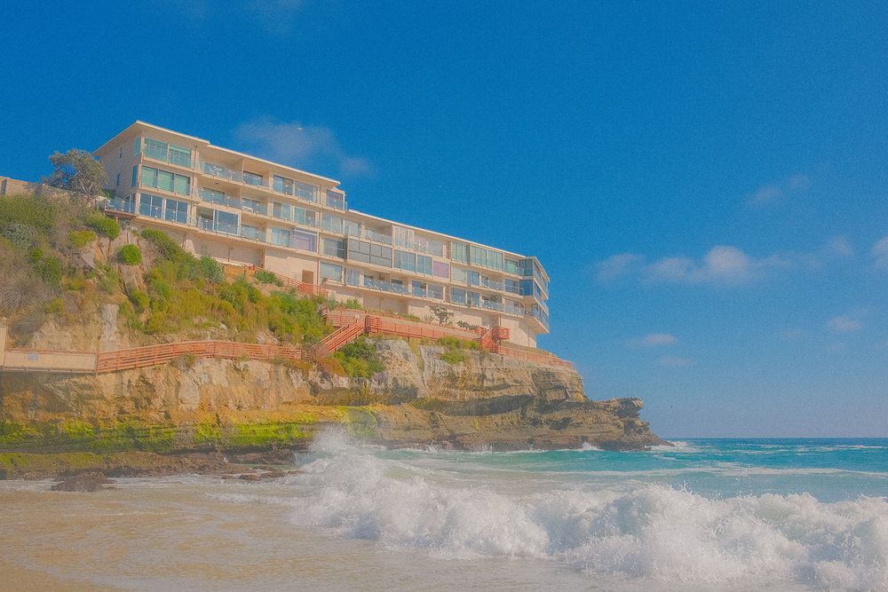 Panoramic view of a beachfront resort hotel with waves crashing against rocky cliffs on a sunny day.