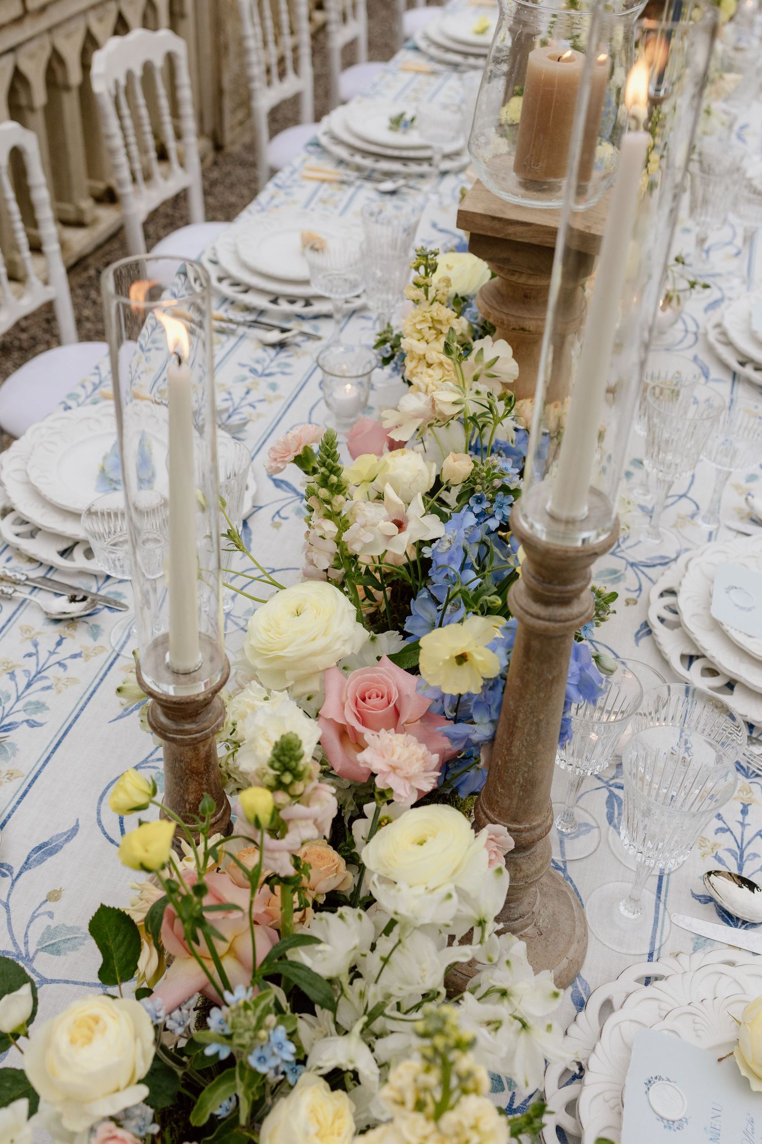 Elegant wedding table setting with delicate blue, pink and white floral centerpieces and tall candlesticks on ornate linens. Destination wed