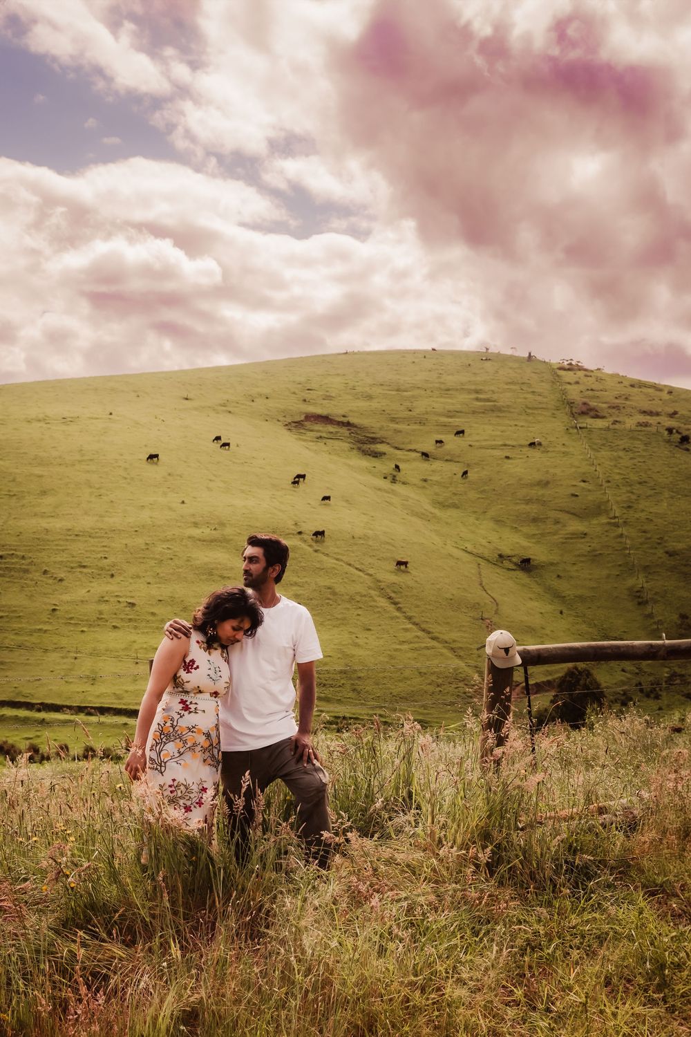 A couple shares a romantic moment on a grassy hillside beneath pink-tinted clouds.