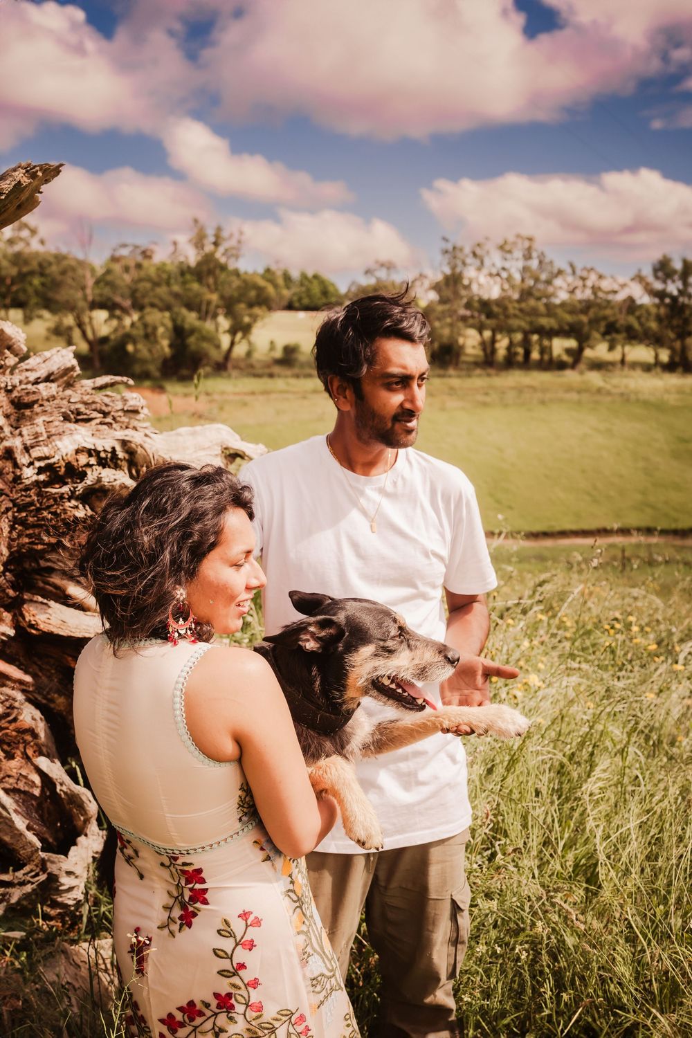 Couple enjoying a sunny day outdoors with their happy dog in a pastoral field with fluffy clouds overhead.