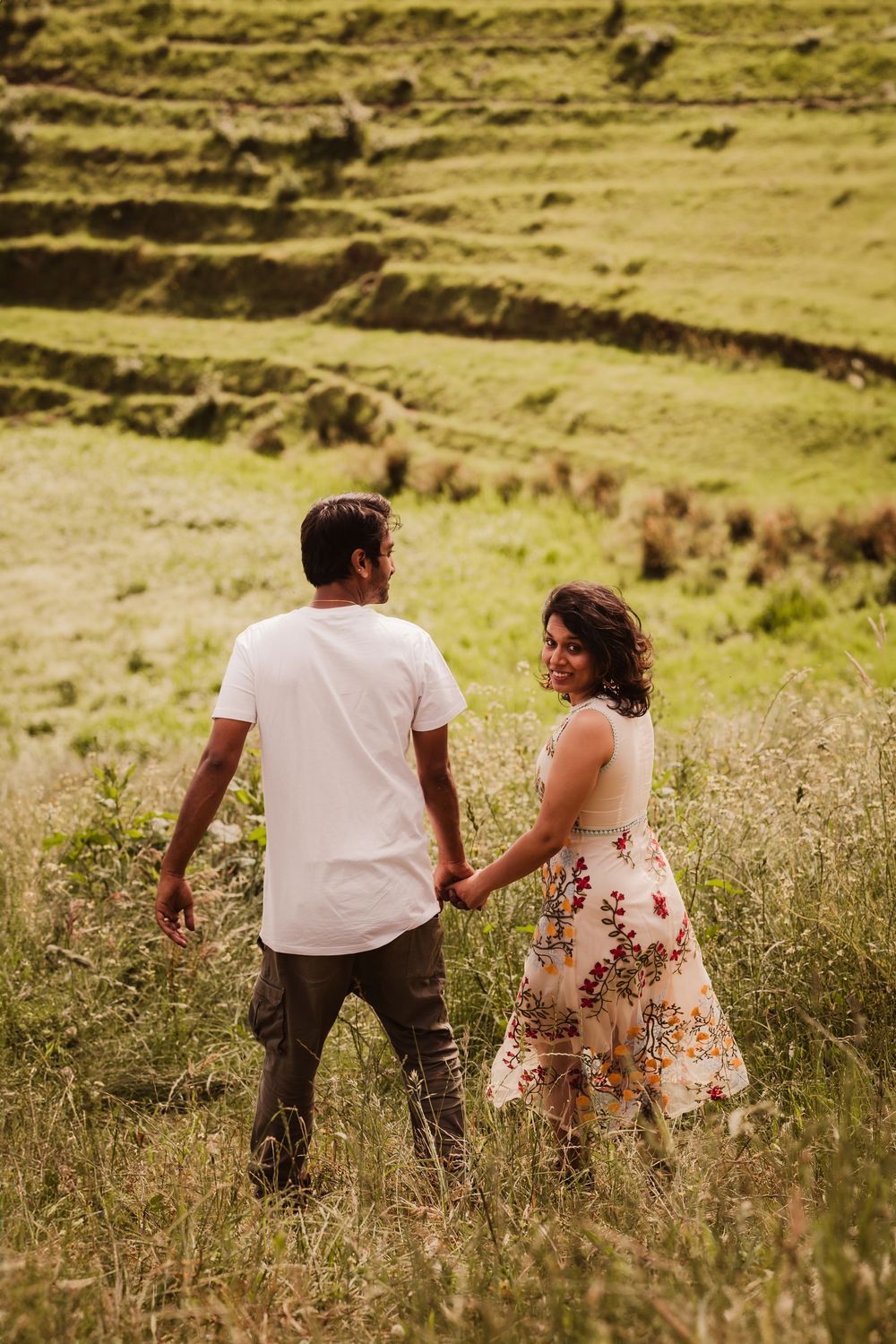 A couple walks hand in hand through a grassy hillside with terraced slopes in the background.