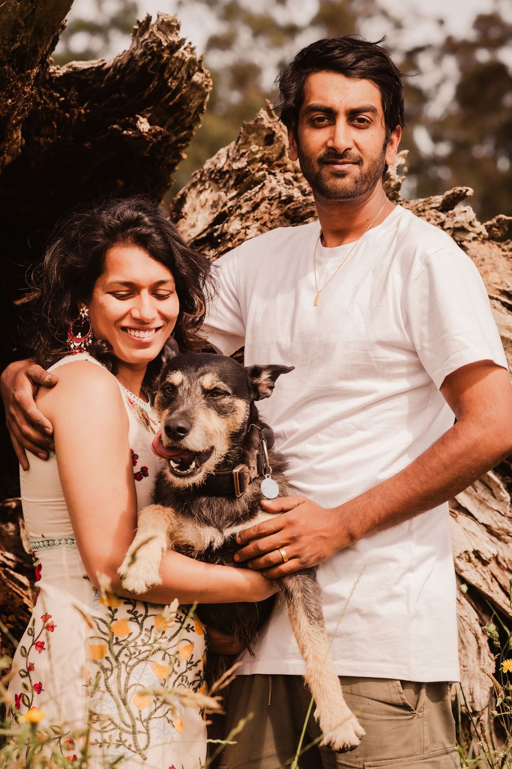 A happy couple in casual attire embraces their scruffy senior dog while posing outdoors against a rustic wooden backdrop.