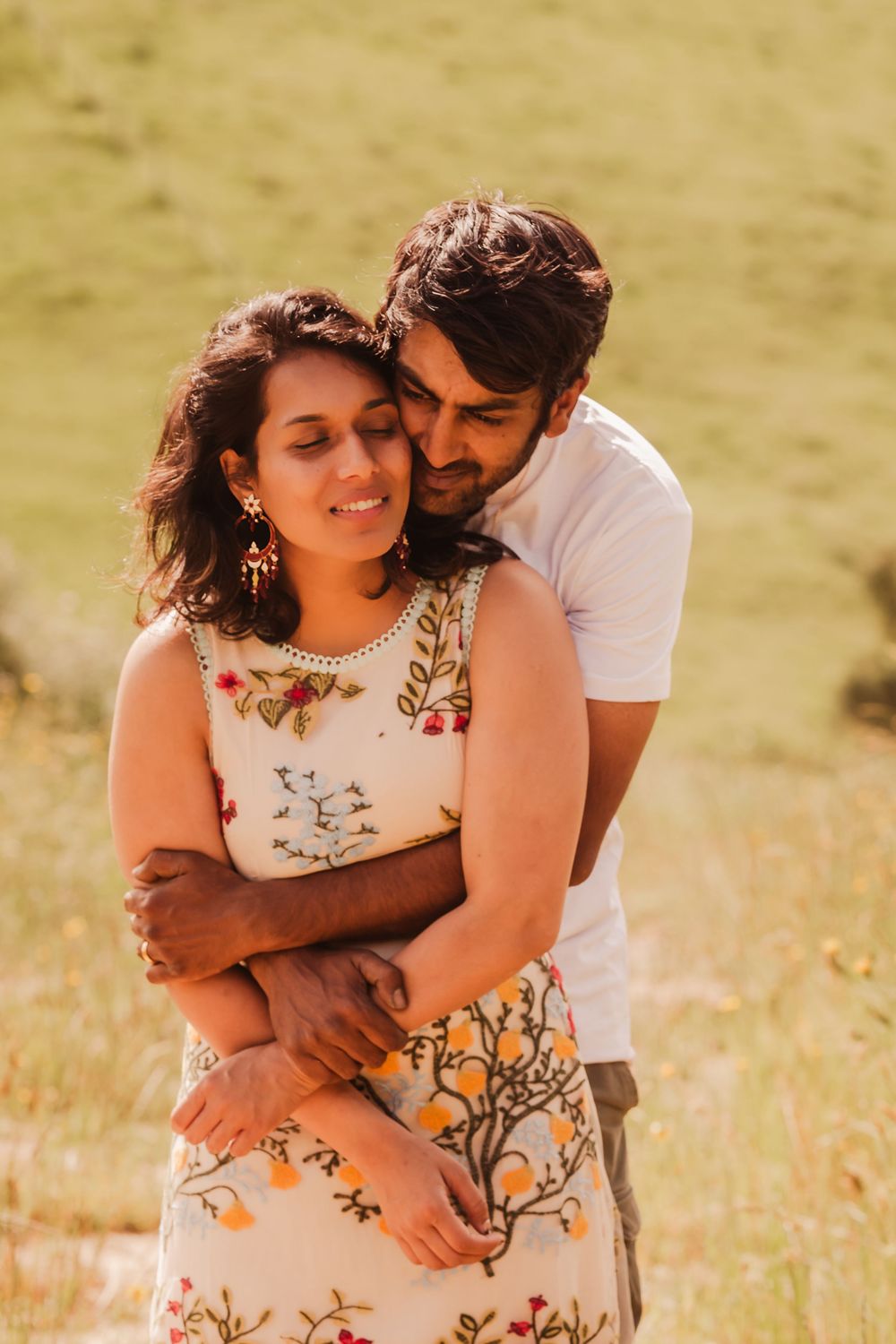 Two people share an intimate embrace in a golden field, wearing casual attire and natural daylight surrounds them.