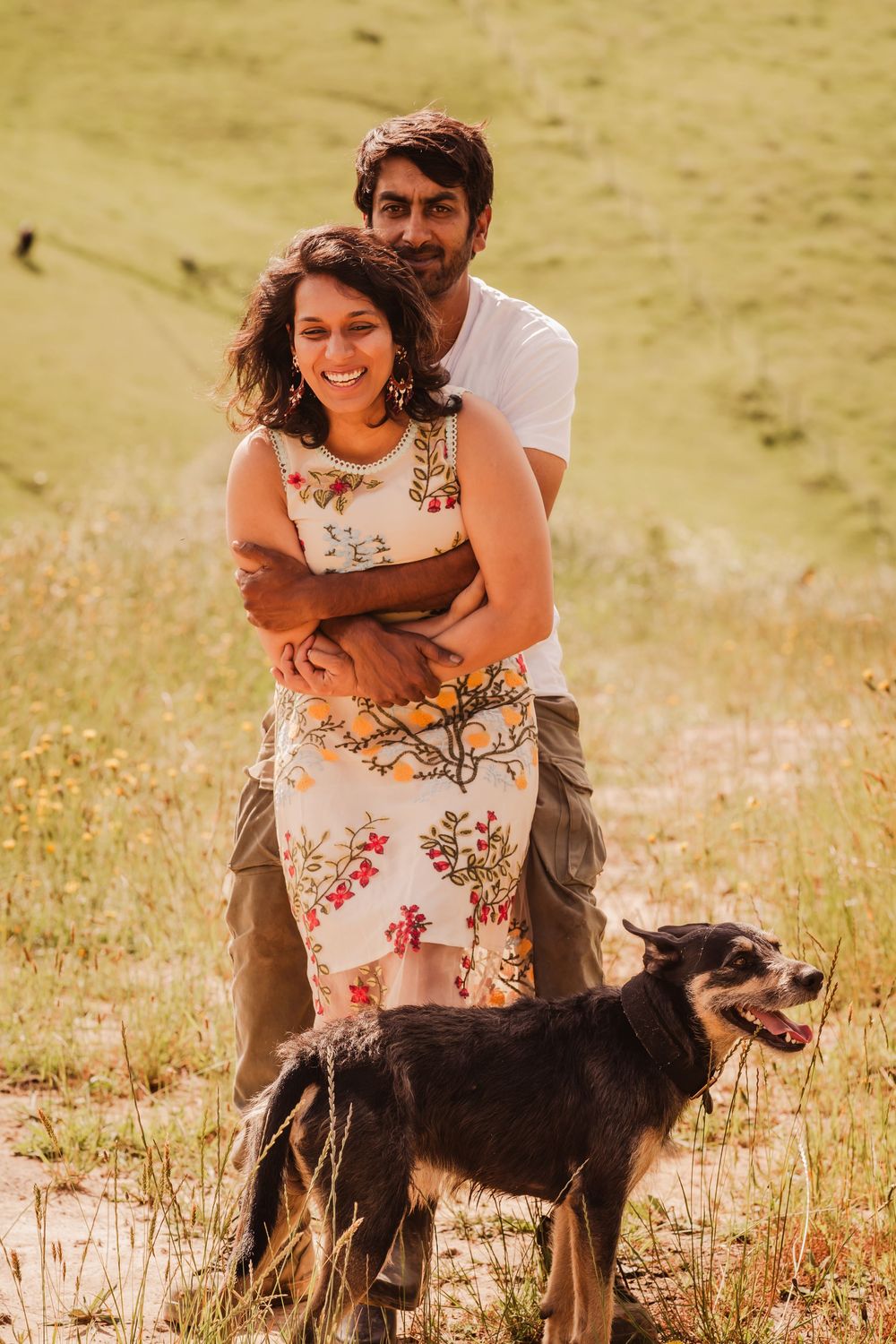 A couple embraces in a sunny meadow field with their black and brown dog standing beside them in warm summer light.