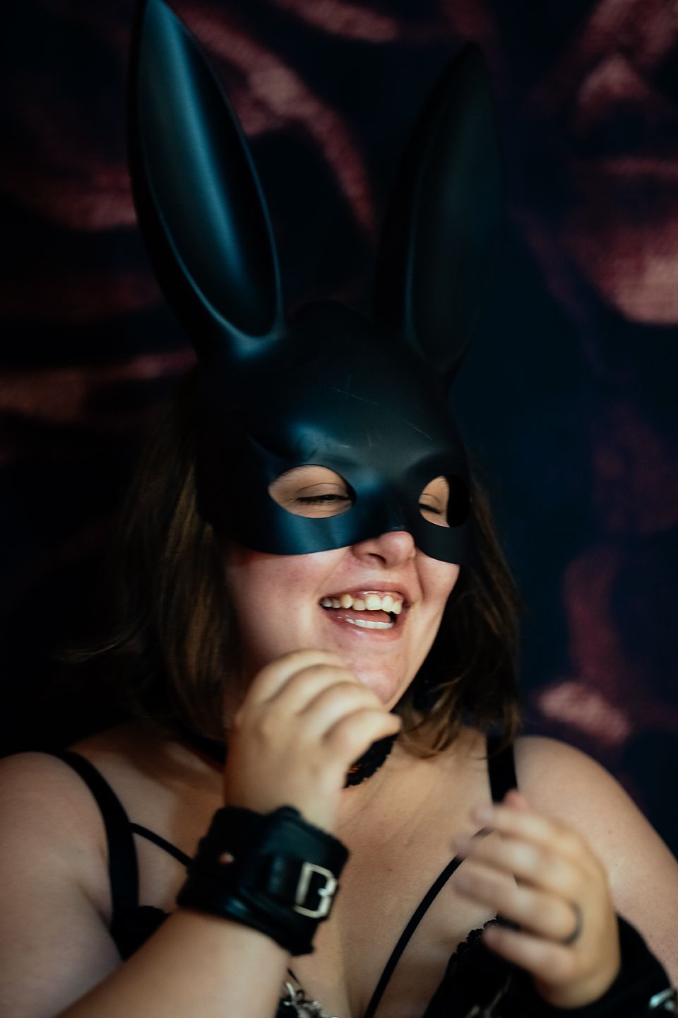 Individual in dark mask and leather collar smiles while in shadows with atmospheric blue lighting.