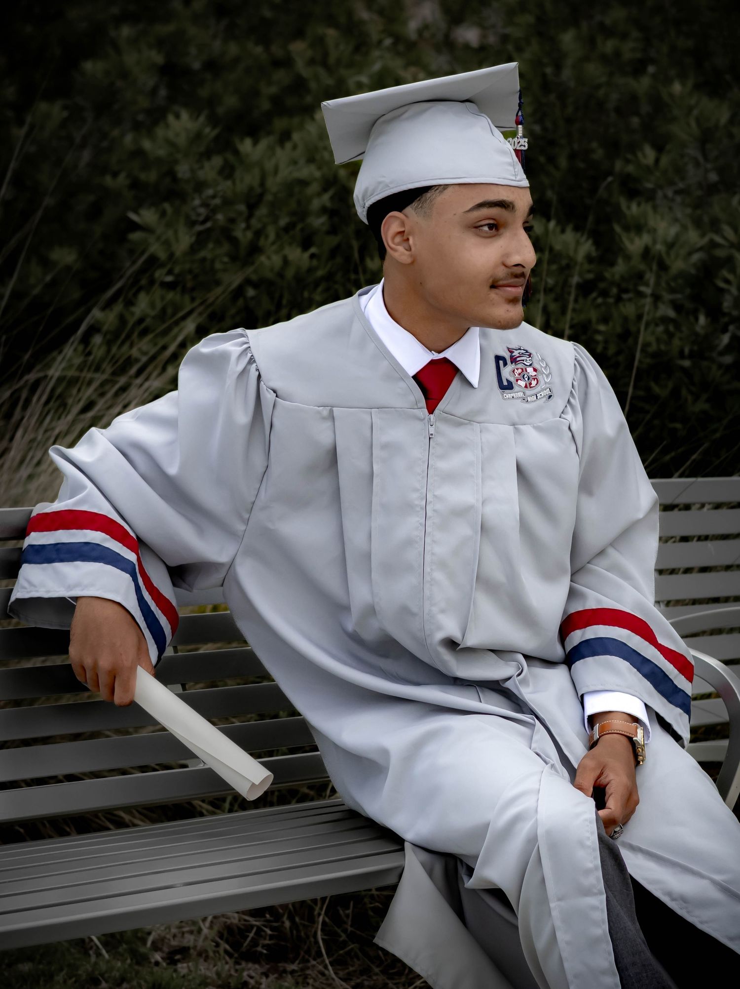 A graduate in white cap and gown with red tie sits on a bench holding a diploma.