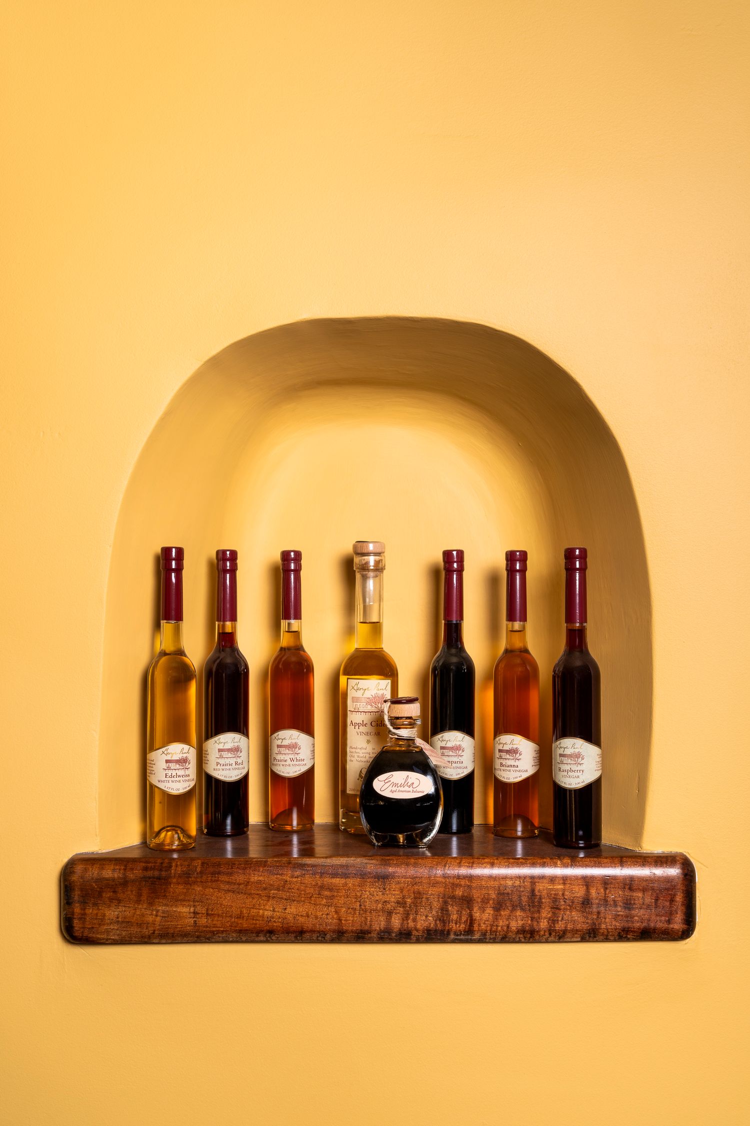 Seven wine bottles displayed on a wooden shelf within a golden-lit alcove niche.