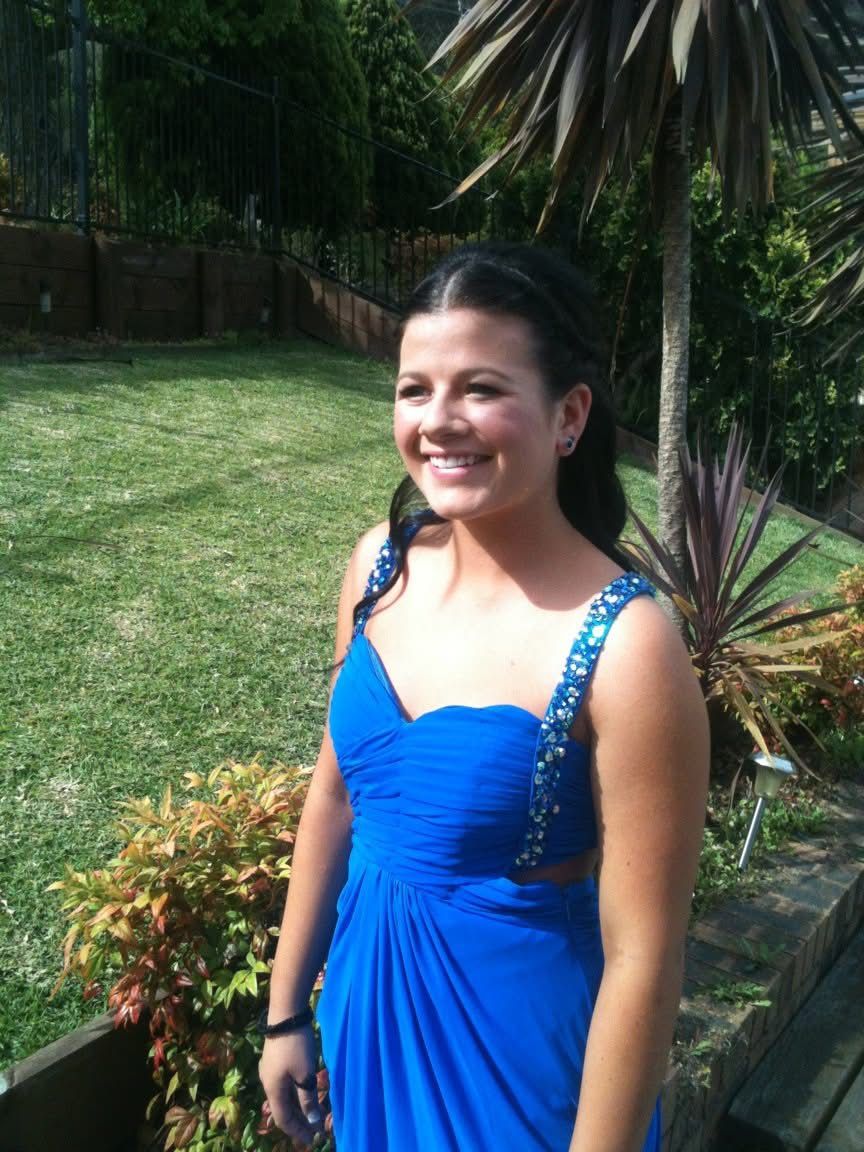 A person poses in a bright blue formal dress with beaded straps while standing in a garden setting.