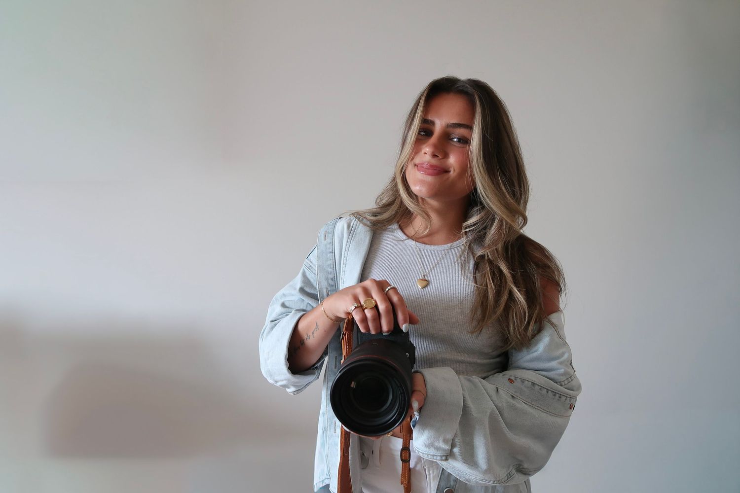 A photographer holding a professional camera wears a light gray sweater against a white wall.