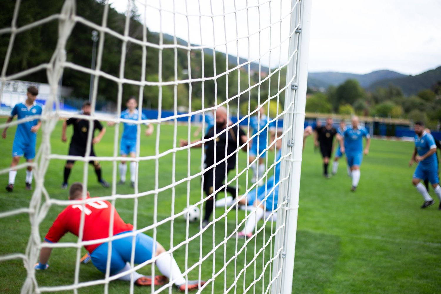 Football players viewed through a goal net during a match on a green pitch in the mountains.