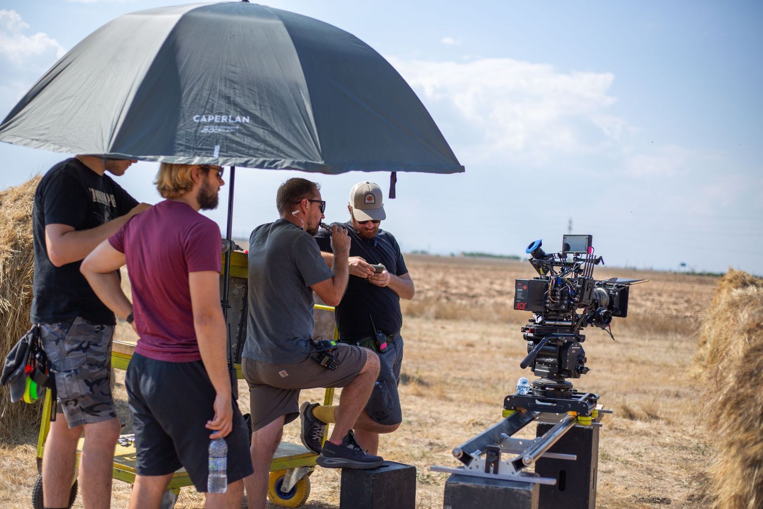 A film crew gathers under an umbrella around a professional camera on a dry open field location.