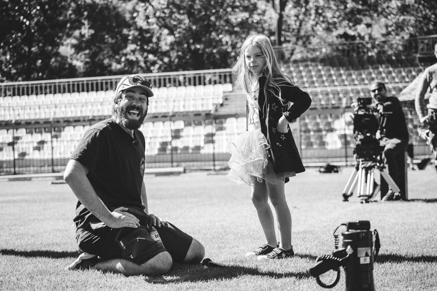 A black and white photo of a cameraman kneeling beside a young girl on a stadium field.