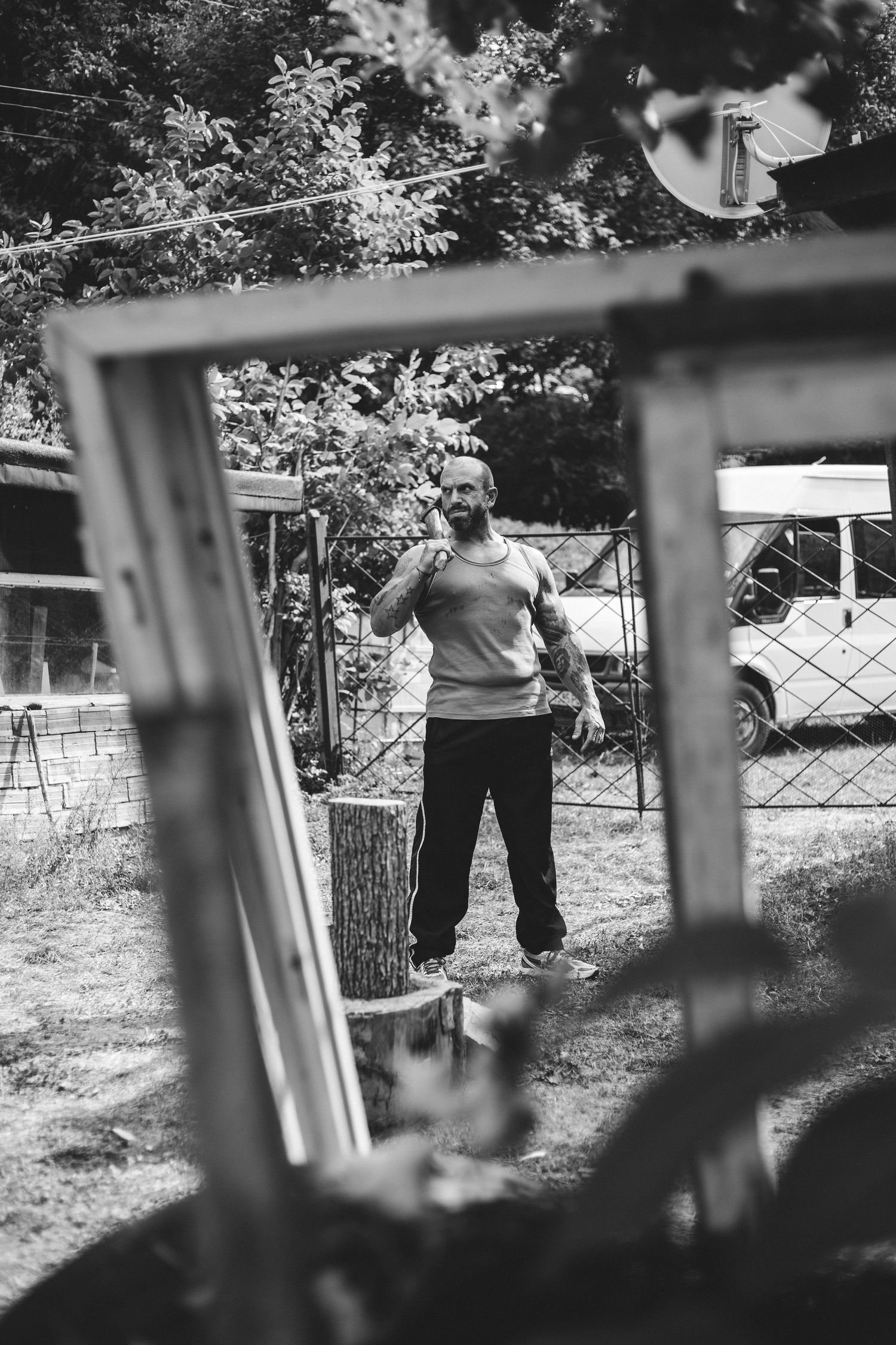 Black and white photo of bald man standing in backyard framed by window.