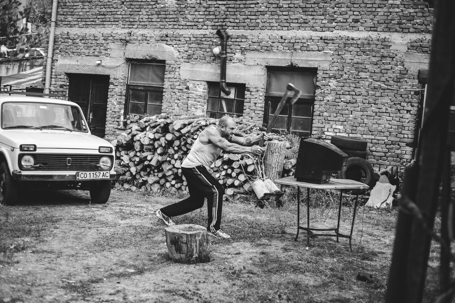 Black and white photo of person carrying firewood in rustic courtyard.