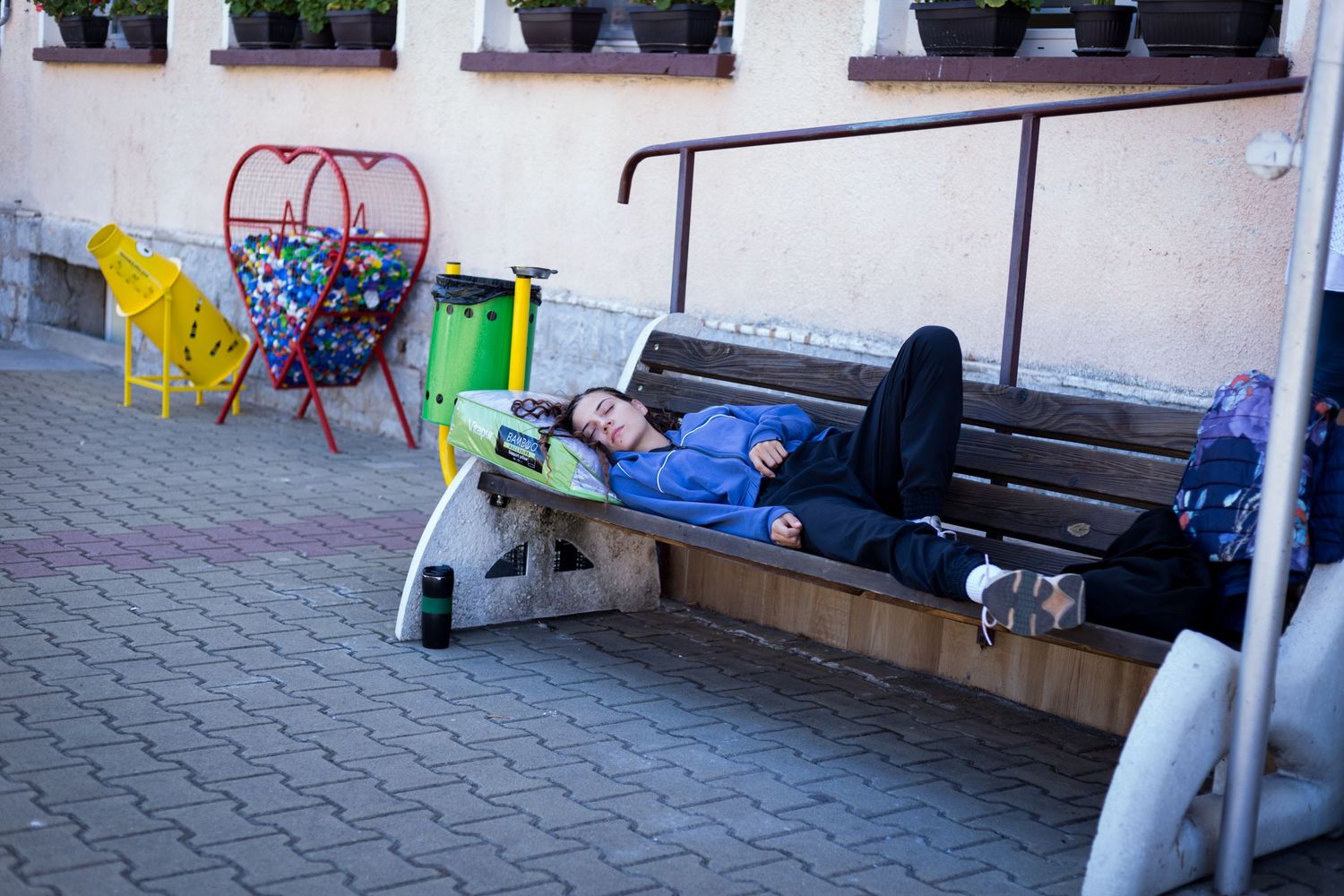 Person lying relaxed on outdoor bench with skateboard nearby in urban pedestrian area with colorful street furniture.