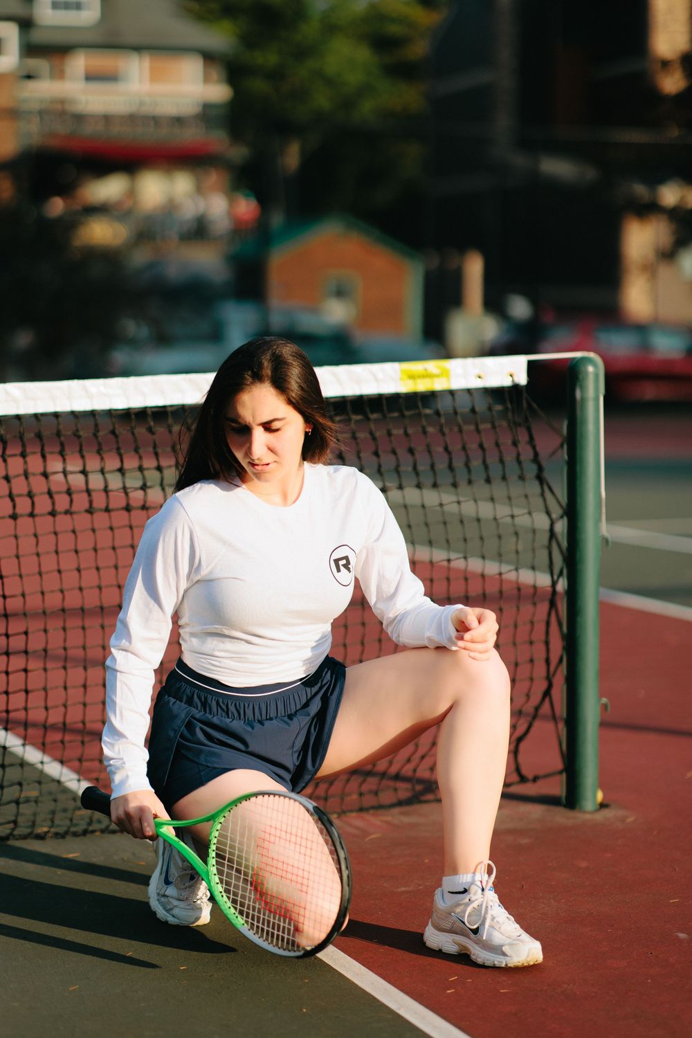 Athletic person kneeling on a tennis court with a racket wearing white long sleeves and shorts.