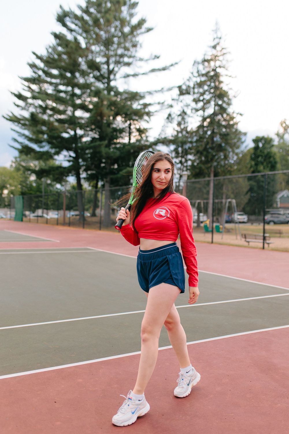 Athlete in red cropped sweatshirt and navy shorts poses on tennis court with evergreen trees in background.
