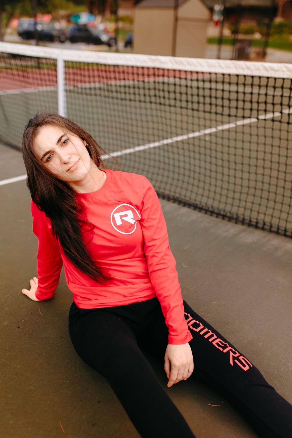 Person wearing red sweatshirt and black pants reclining along tennis court net.