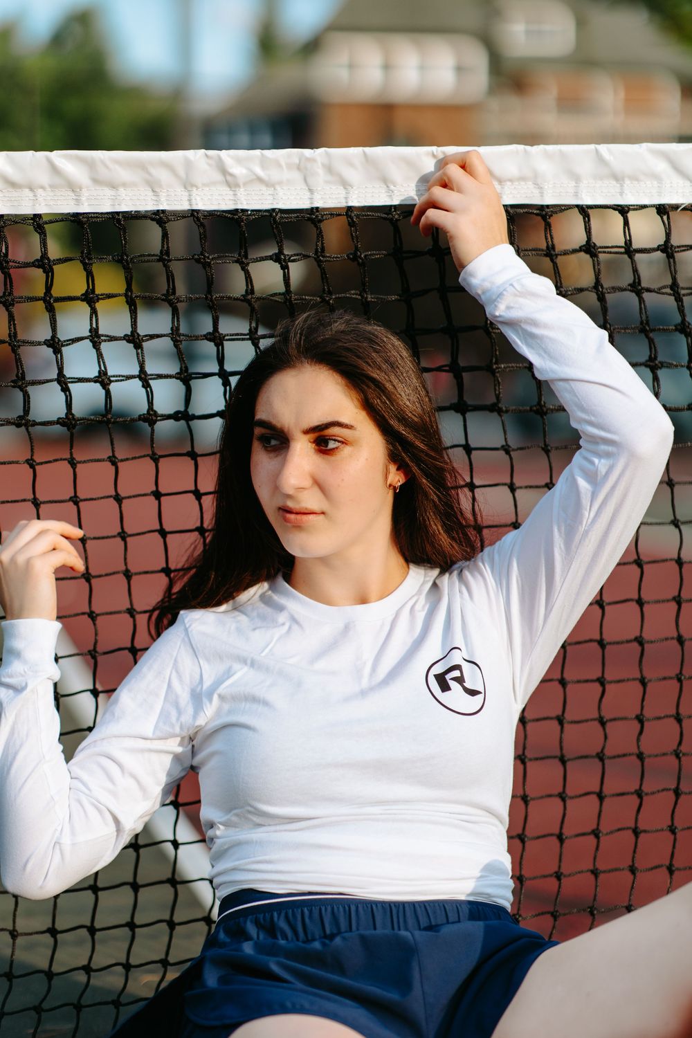 Athletic model poses by tennis court net wearing white long sleeve top and navy shorts.