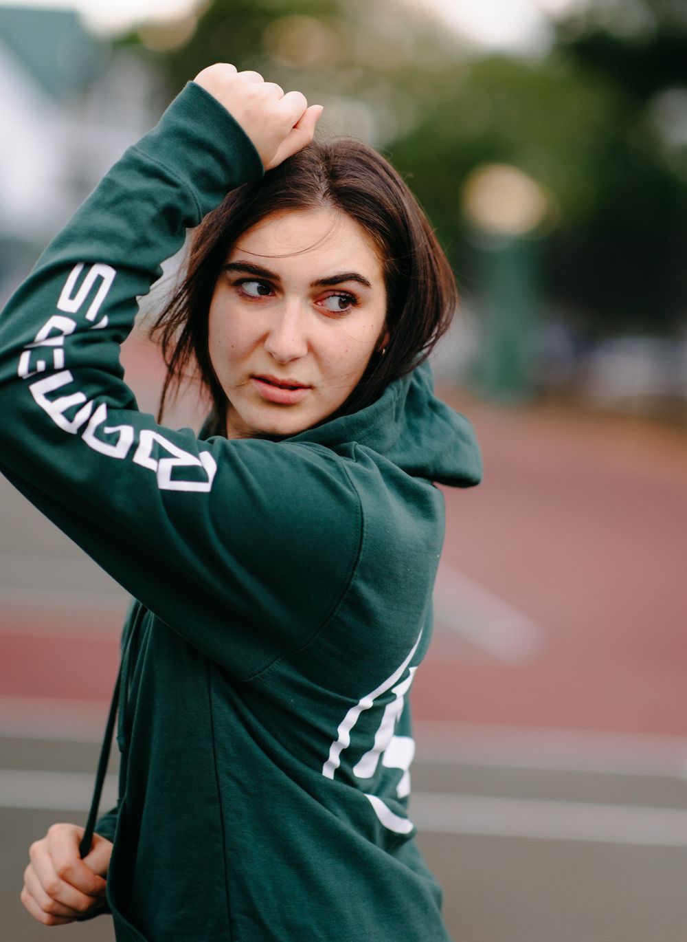 Person wearing dark green athletic hoodie with white text branding posing outdoors at a sports track.