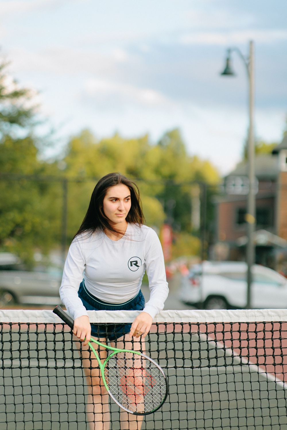 Athletic person in white shirt leaning over tennis court net on outdoor court with trees in background.