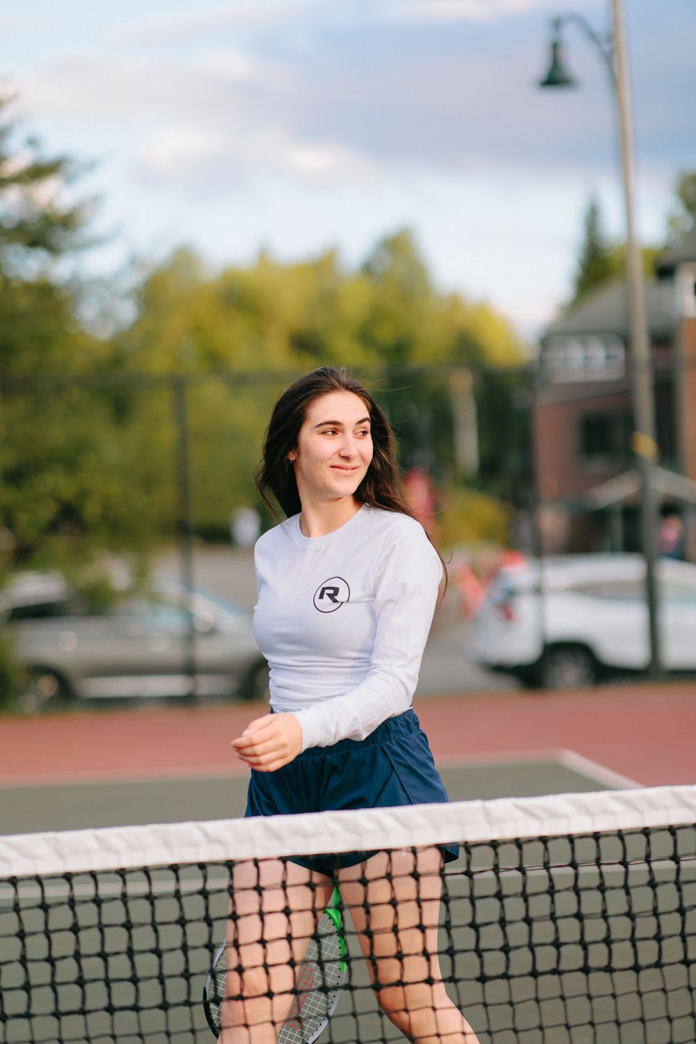 Person in casual attire posing playfully along tennis net on outdoor court.