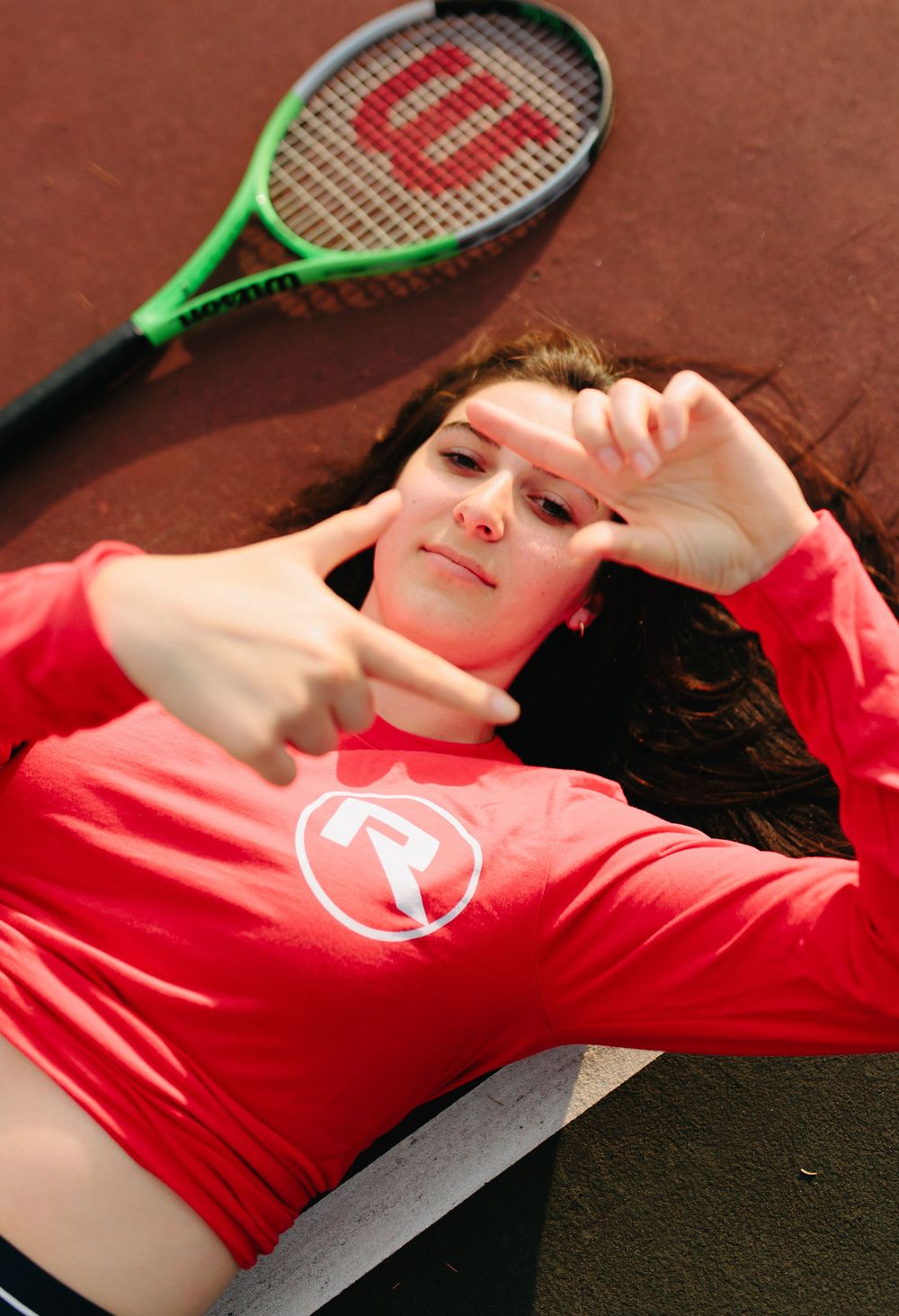 A person in a red shirt lies back on a dark surface with tennis rackets visible in the background.