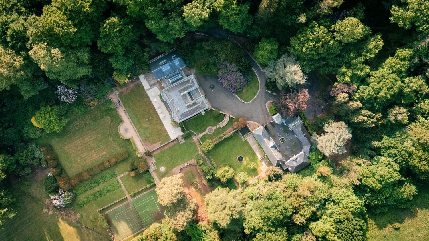 Aerial drone view of residential property with tennis court and landscaped gardens surrounded by lush green trees.