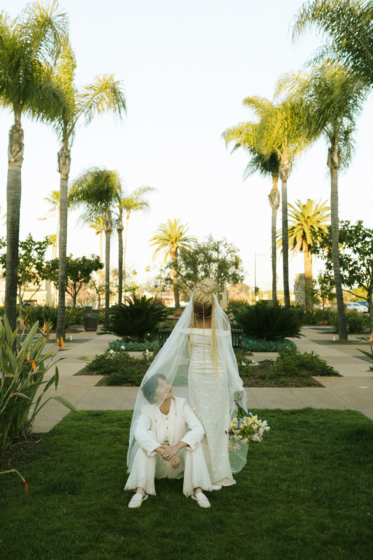 One bride sits on the grass in front of Laguna Hills CIty Center while their wife stands over them, draping their veil around them both