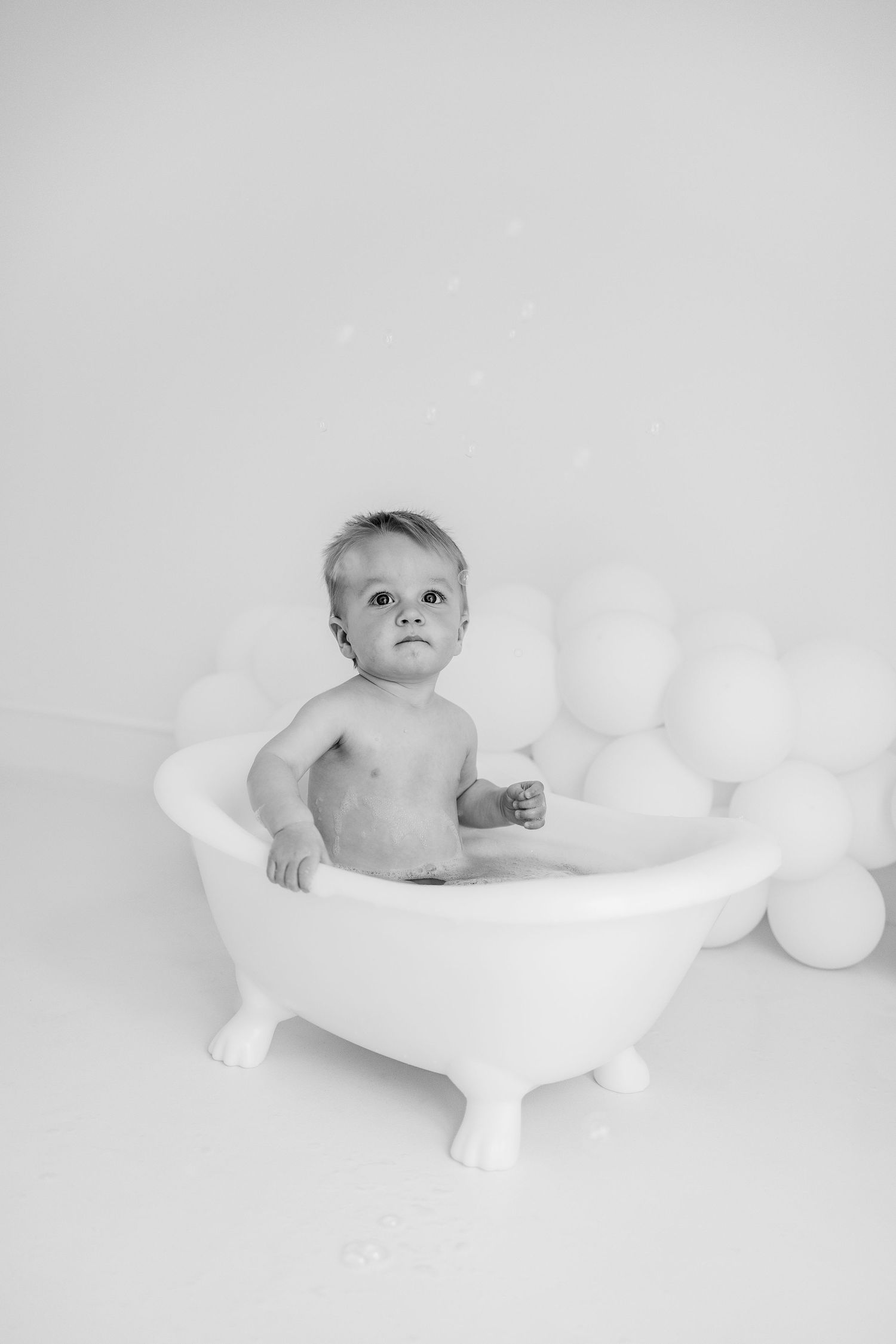 A small baby sits in a classic white clawfoot bathtub surrounded by white balloons in a minimalist black and white photo.