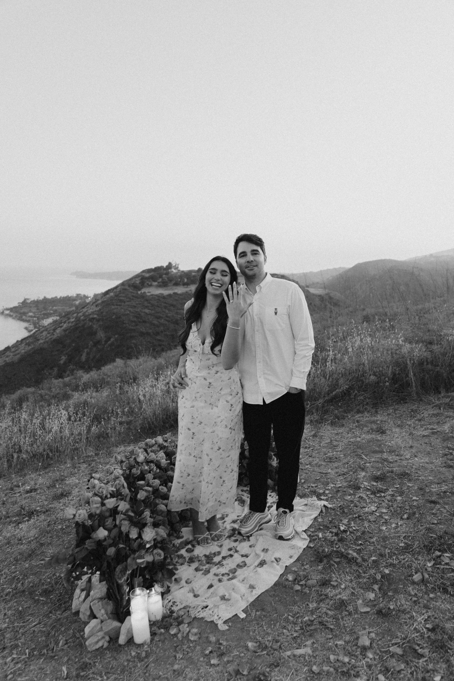A couple stands on a hilltop overlooking the coast at sunset with candles and flowers placed at their feet.