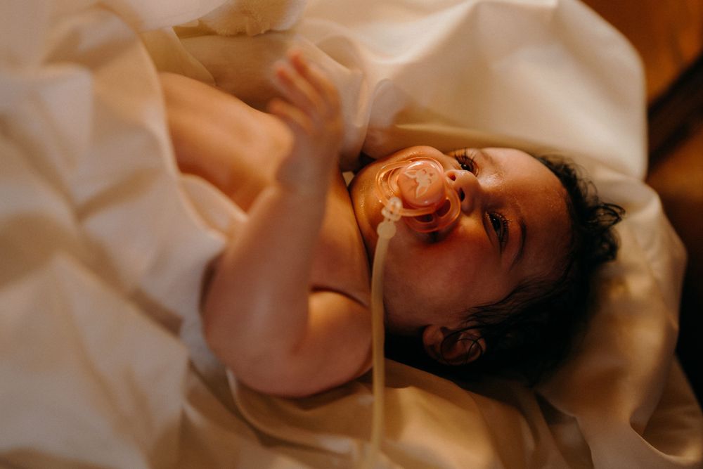 A baptized Melbourne baby wrapped in a soft white blanket smiles peacefully with a pacifier, starring at his mother under a golden sunlight.