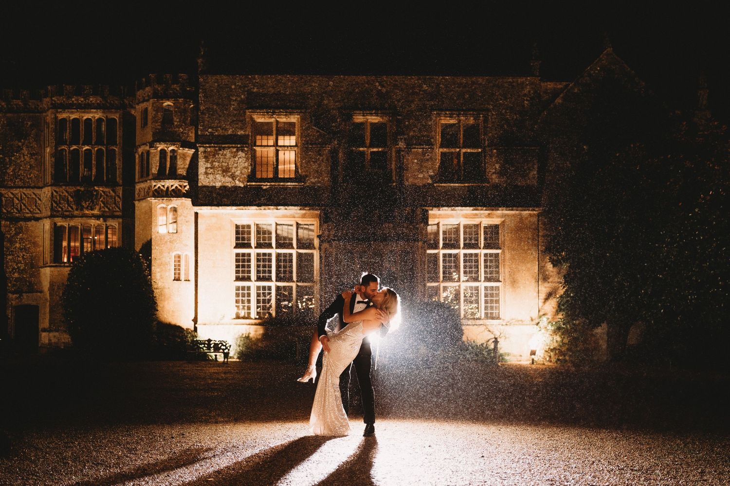 Two silhouettes at a wedding in a photo embrace in dramatic backlit scene outside illuminated historic stone building at night.