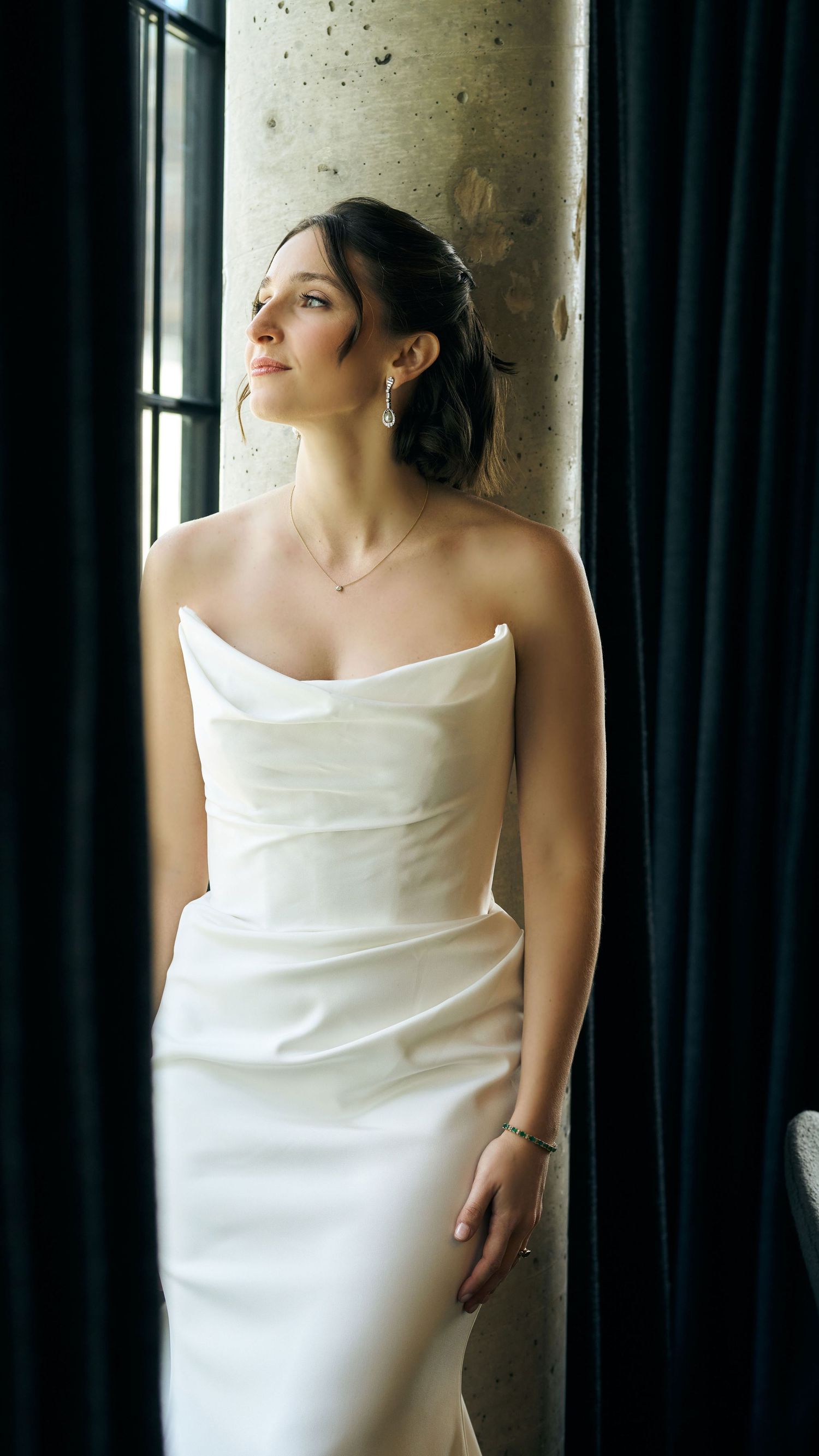 Elegant bridal portrait showing a white strapless wedding gown against dark curtains near a window.