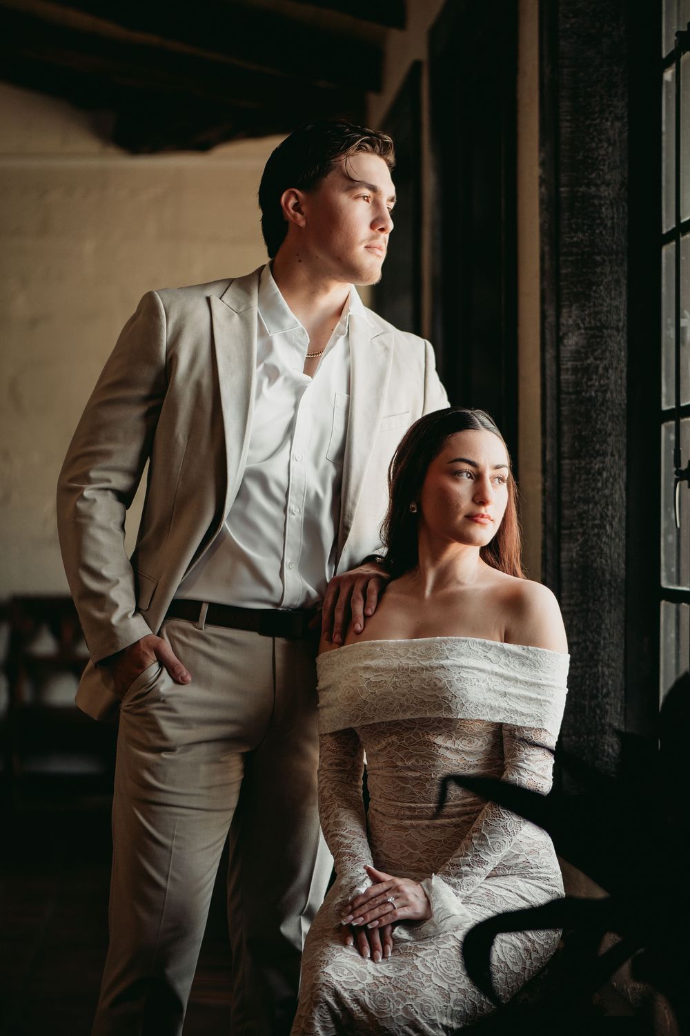 A couple poses together in a moody indoor setting with dramatic window lighting and vintage aesthetics.
