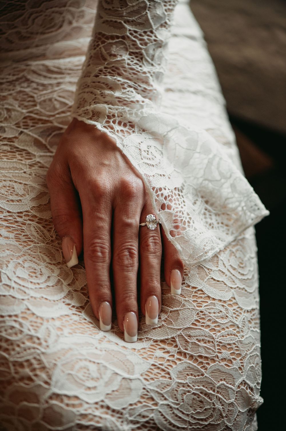 Close-up view of a hand with manicured nails resting on white lacy wedding dress fabric, showcasing an engagement ring.