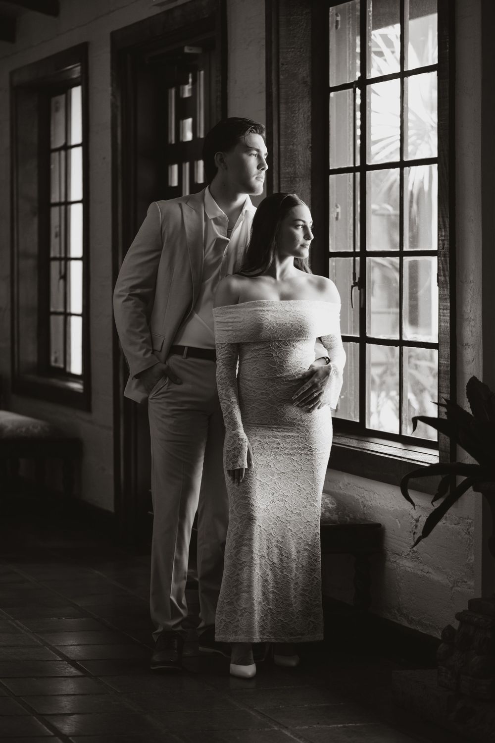 A black and white romantic portrait of a couple standing by large windows in formal attire.