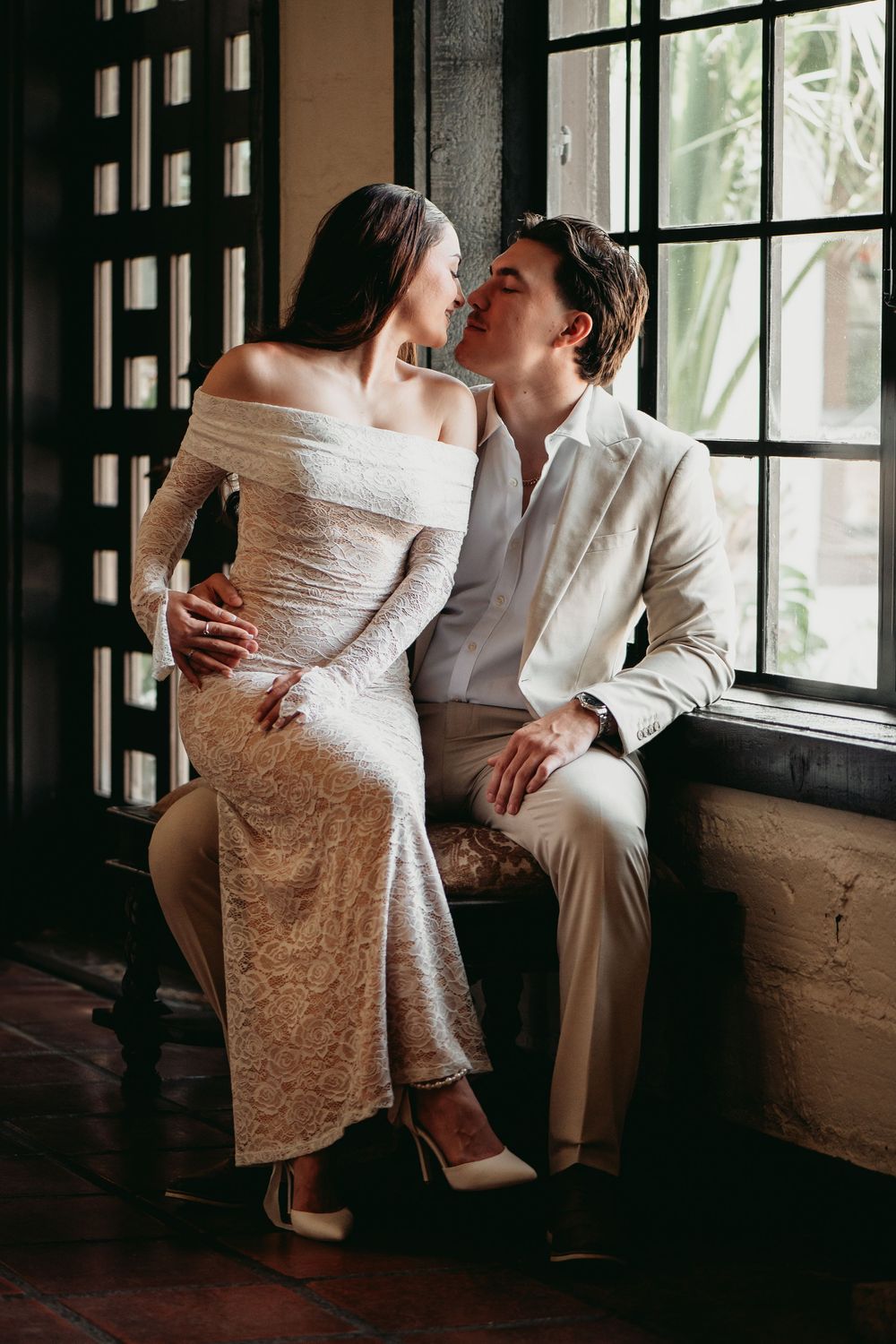 A romantic couple shares an intimate moment by a window in an artistic black and white photo series.