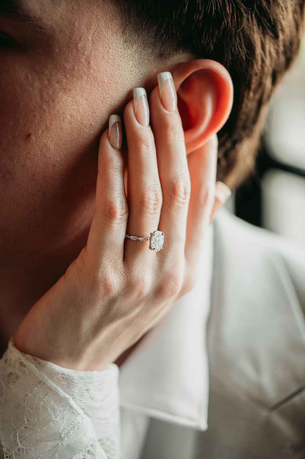 Close-up of a hand with white lace sleeve displaying an elegant diamond engagement ring next to an ear.