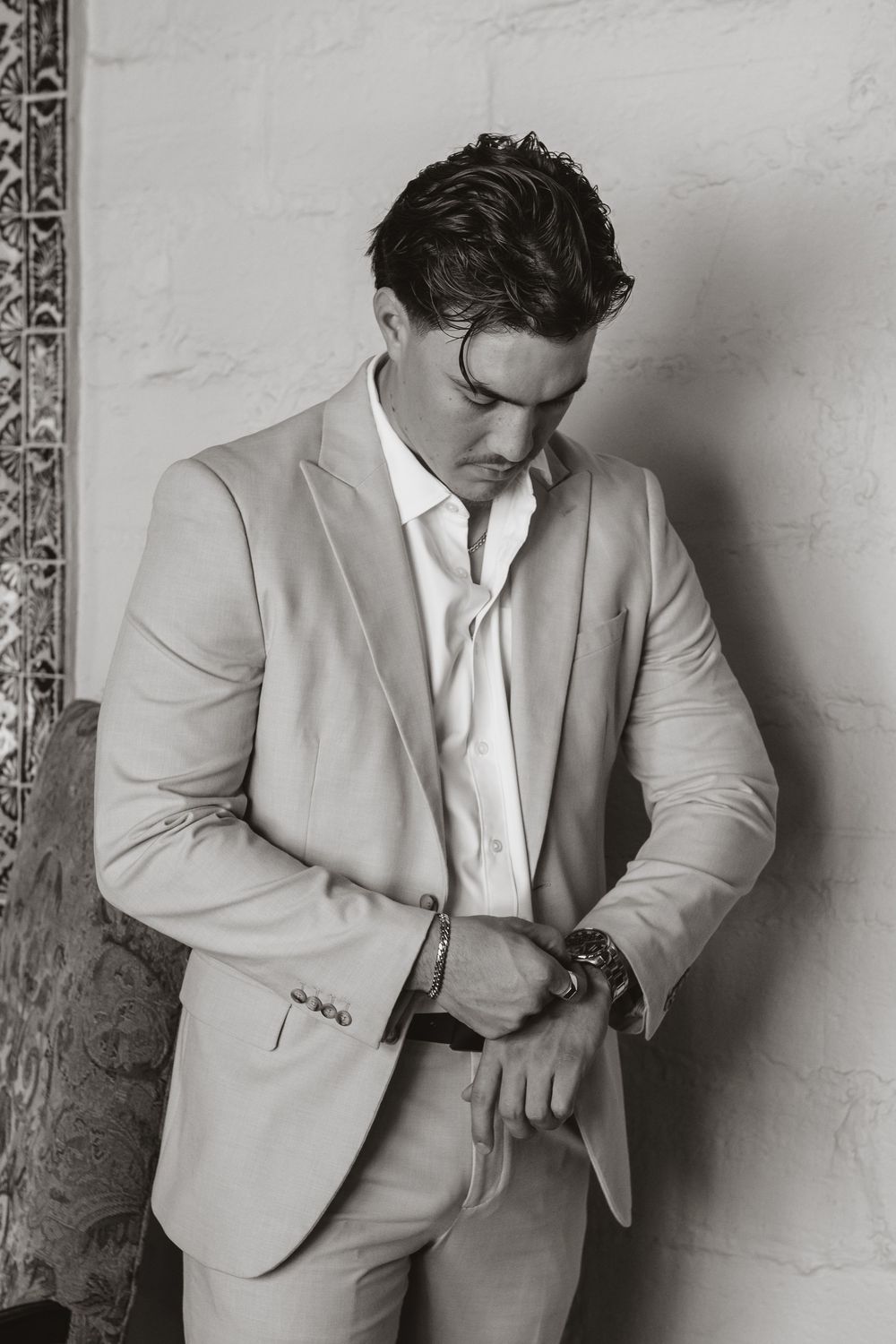 A black and white portrait of someone in a light colored suit checking their watch against a textured wall.
