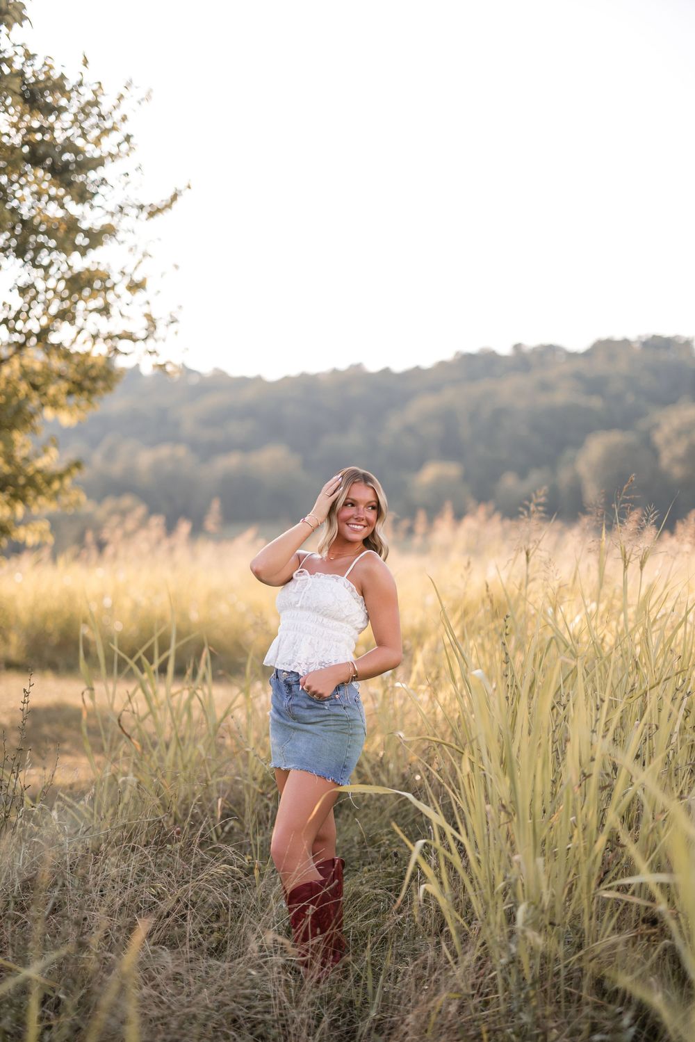 A woman stands in tall grass at sunset wearing a white top, denim skirt and brown cowboy boots in a rural setting.