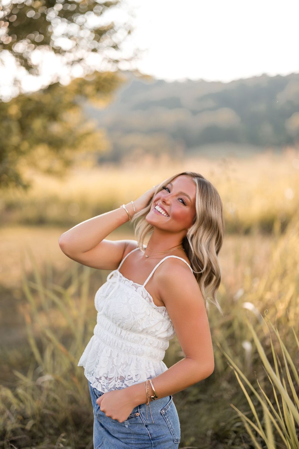Woman wearing white lace top and jeans poses joyfully in a sunny field at golden hour.